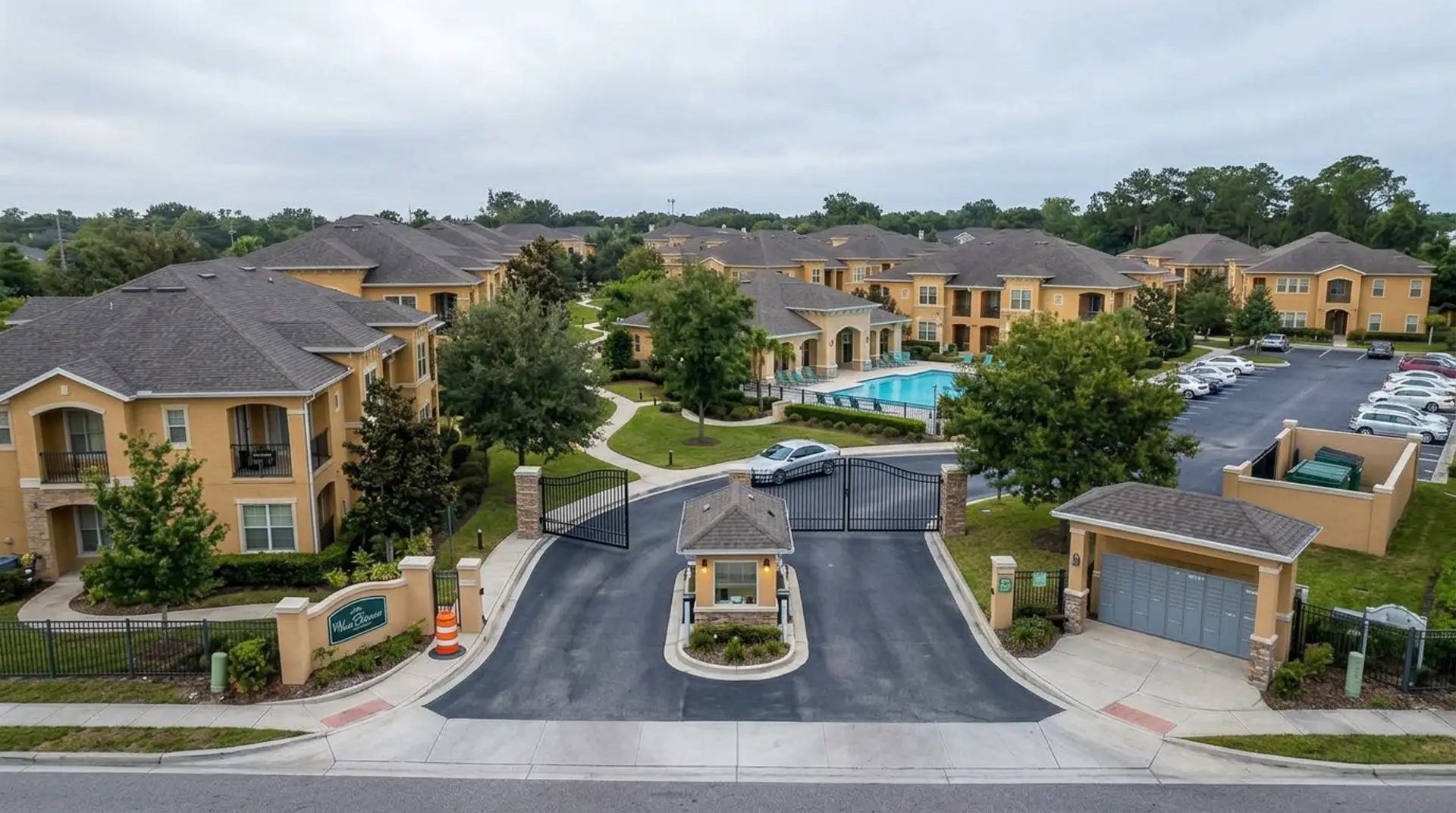 An upscale gated community entrance on an overcast day: one metal swing gate standing wide open with a repair barrel beside it while a car drives through unchallenged